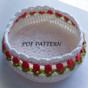 Peut inclure: Un panier crocheté blanc avec un bord décoratif orné de motifs floraux rouges et verts. Le panier est de forme ronde et le texte "PDF PATTERN" est visible sur le côté.