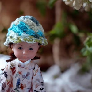 Peut inclure: Une poupée portant un chapeau crocheté à la main avec du fil bleu, vert et crème. La poupée porte une robe à imprimé floral. Le chapeau a un bord et est crocheté dans un motif rayé. La poupée a la peau pâle et des cheveux bruns.