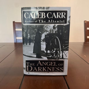 Op de afbeelding: Een hardcover boek getiteld "The Angel of Darkness" van Caleb Carr, auteur van "The Alienist". De omslag toont een zwart-wit afbeelding van een paardenkoets en figuren op een straat.