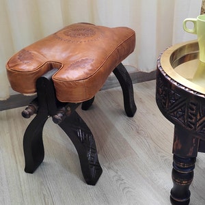 May include: A brown leather ottoman with a carved wooden base and a decorative brass tray table with carved wooden legs. The tray table has a green ceramic mug and a black ceramic cup on it.