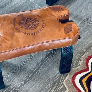 May include: A brown leather bench with intricate carved designs on the seat and legs. The bench is made of wood and has a rustic look.