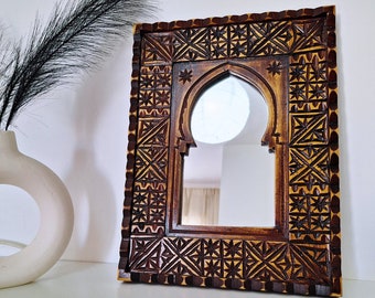 May include: A wooden mirror with a carved, intricate design in shades of brown and gold. The mirror has an arched top and a rectangular frame.