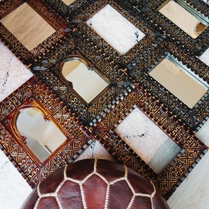 May include: A set of intricately carved wooden frames with ornate designs. The frames are arranged in a staggered pattern and feature a variety of sizes and shapes. The frames are made of dark wood with a golden finish. A brown leather pouf sits in the foreground.