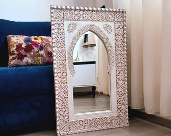 Op de afbeelding: Een witte houten spiegel met een ingewikkeld gesneden ontwerp. De spiegel heeft een gebogen bovenkant en is omlijst door een rand met geometrische patronen. De spiegel leunt tegen een blauwe bank.