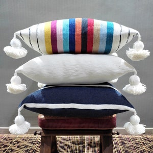 Peut inclure: Trois coussins décoratifs avec des pompons blancs empilés sur un tabouret en bois avec un siège bordeaux. Le coussin du haut est à rayures dans des tons de rose, jaune, bleu et blanc. Le coussin du milieu est blanc et le coussin du bas est bleu marine à rayures blanches.