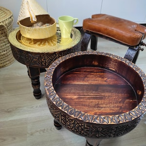 Peut inclure: Deux tables en bois sculptées avec des plateaux ronds. La table de gauche a un plateau en laiton avec un bol décoratif en or. La table de droite a un plateau en bois brun foncé.