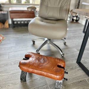 Op de afbeelding: Een bruine leren voetenbank met ingewikkelde houtsnijwerk en een decoratief ontwerp. De voetenbank is gemaakt van hout en heeft een metalen basis.