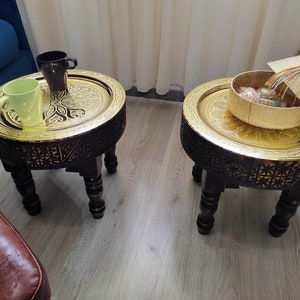 May include: Two round, gold-colored, decorative trays with intricate carvings on the sides, each sitting on a dark wood stand with three legs. The trays are on a light wood floor.
