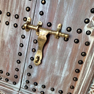 Könnte beinhalten: Nahaufnahme eines Türverschlusses aus Messing mit einem verzierten Design, montiert an einer weiß getünchten Holztür mit dekorativen Metallnieten.