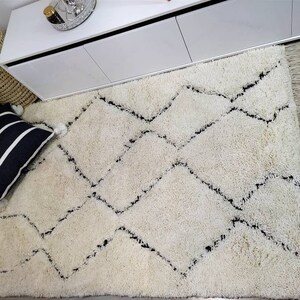 May include: A white shag rug with a black diamond pattern. The rug has a fringe border and is laid out on a hardwood floor.