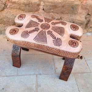 May include: A traditional Moroccan leather pouf with intricate carved wooden legs. The pouf is decorated with a geometric pattern and has a light brown color.