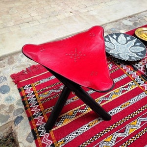 May include: A red leather folding stool with black wooden legs. The stool is sitting on a red and black patterned rug.