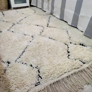 May include: A white shag rug with a black diamond pattern. The rug has a fringe border and is laid out on a hardwood floor.