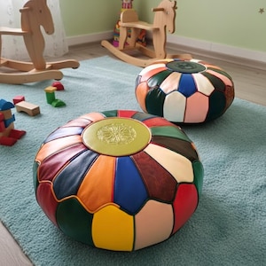 Könnte beinhalten: Zwei runde, bunte Leder-Puffs mit Patchwork-Design. Die Puffs befinden sich in einem Kinderspielzimmer, mit Holzschaukelpferden und Spielzeug im Hintergrund. Die Puffs haben eine Vielzahl von Farben, darunter Rot, Blau, Grün und Gelb.