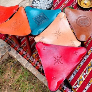 May include: Five triangular leather poufs with intricate geometric designs. The poufs are in shades of red, orange, blue, brown, and pink. They are arranged on a colorful rug with a geometric pattern.