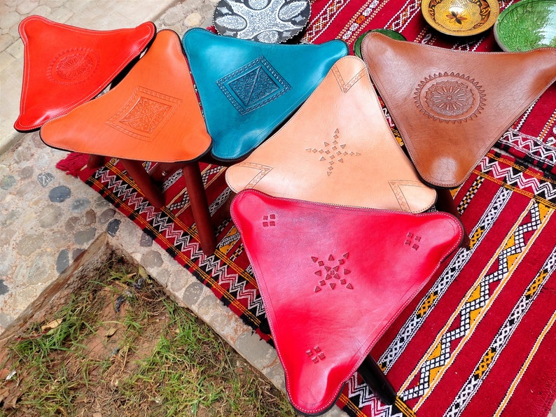 May include: Five triangular leather poufs with intricate geometric designs. The poufs are in shades of red, orange, blue, brown, and pink. They are arranged on a colorful rug with a geometric pattern.