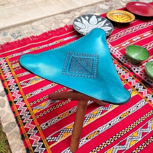 Op de afbeelding: Een turquoise leren poef met een decoratief reli&euml;fpatroon staat op een rood en zwart gestreept tapijt. De poef heeft een driehoekige vorm en drie houten poten.