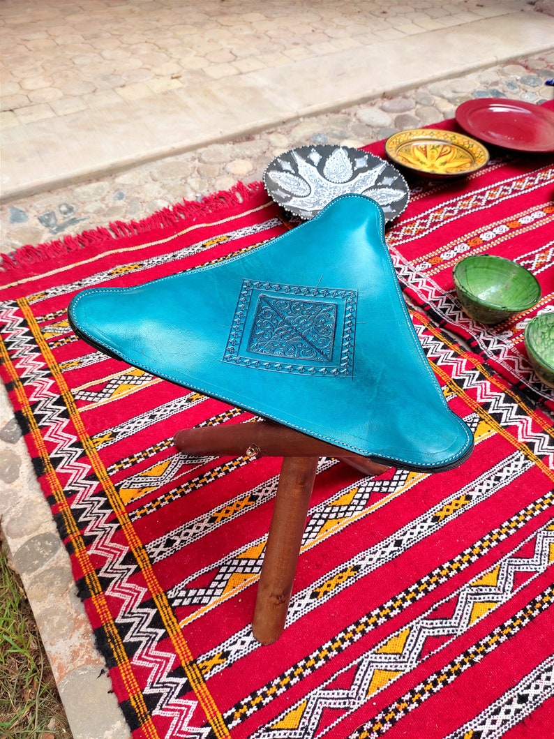Op de afbeelding: Een turquoise leren poef met een decoratief reli&euml;fpatroon staat op een rood en zwart gestreept tapijt. De poef heeft een driehoekige vorm en drie houten poten.