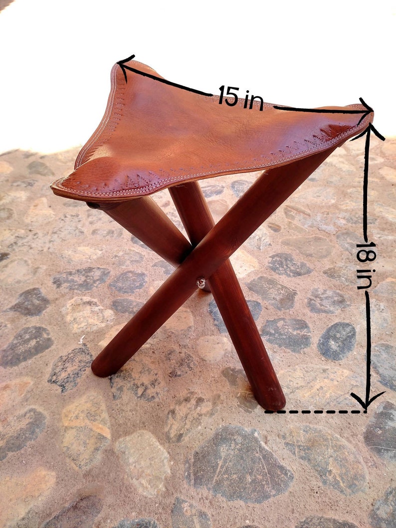 May include: A brown leather folding stool with a wooden frame. The seat is 15 inches wide and the stool is 18 inches tall.