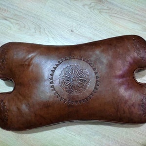 May include: A brown leather pillow with a decorative embossed design in the center. The pillow is shaped like a teardrop and has a rounded edge.