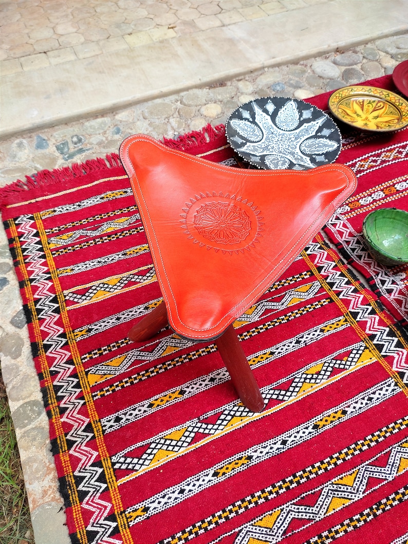 Op de afbeelding: Een rode leren poef met een decoratief ontwerp op de bovenkant. De poef staat op een rood-wit gestreept tapijt.