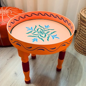 May include: A small orange table with a round top and three legs. The top is painted with a floral design in blue, green, and white. The legs are painted red and orange.