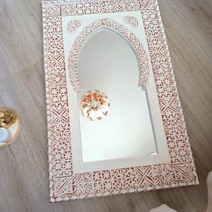 May include: White wooden mirror with a carved, intricate design around the frame. The mirror has an arched top and a white background.