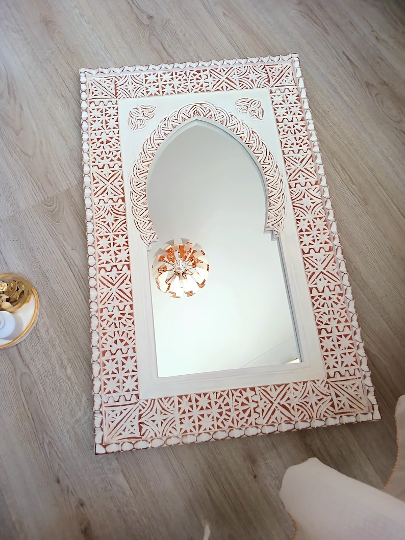 May include: White wooden mirror with a carved, intricate design around the frame. The mirror has an arched top and a white background.