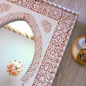 May include: White wooden mirror with an ornate, carved frame. The frame features a repeating geometric pattern in shades of red and white. The mirror has an arched top and a clear reflection.