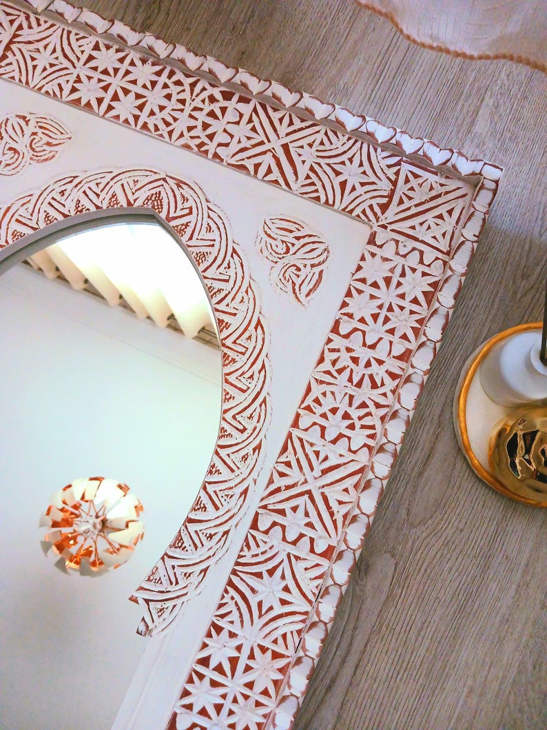 May include: White wooden mirror with an ornate, carved frame. The frame features a repeating geometric pattern in shades of red and white. The mirror has an arched top and a clear reflection.