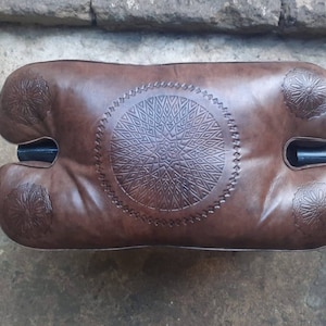 May include: A brown leather pouf with intricate geometric designs embossed on the surface. The pouf has a rounded shape with two separate sections and a black metal frame.