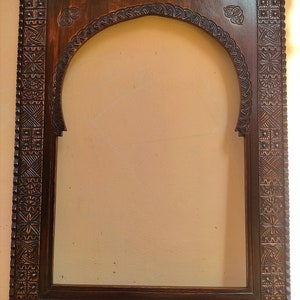 May include: A dark brown, intricately carved wooden frame with an arched top. The frame features detailed geometric and floral patterns, creating a decorative border. The frame is designed to hold a mirror or artwork.