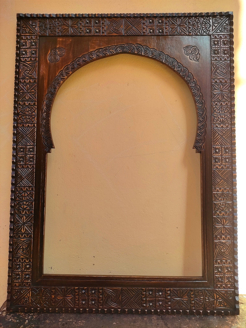 May include: A dark brown, intricately carved wooden frame with an arched top. The frame features detailed geometric and floral patterns, creating a decorative border. The frame is designed to hold a mirror or artwork.