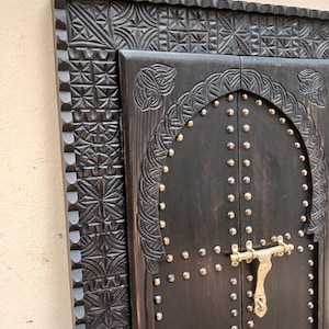 Könnte beinhalten: Ein schwarzer, mit Schnitzereien verzierter Holzrahmenspiegel mit einem dekorativen Türdesign. Die Tür hat einen Messingriegel und ist mit Metallnieten besetzt.