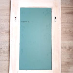 May include: White wooden frame with a light blue mirror inside. The frame has two small white hooks on the back. The mirror has the text "F1 0781" printed on it.