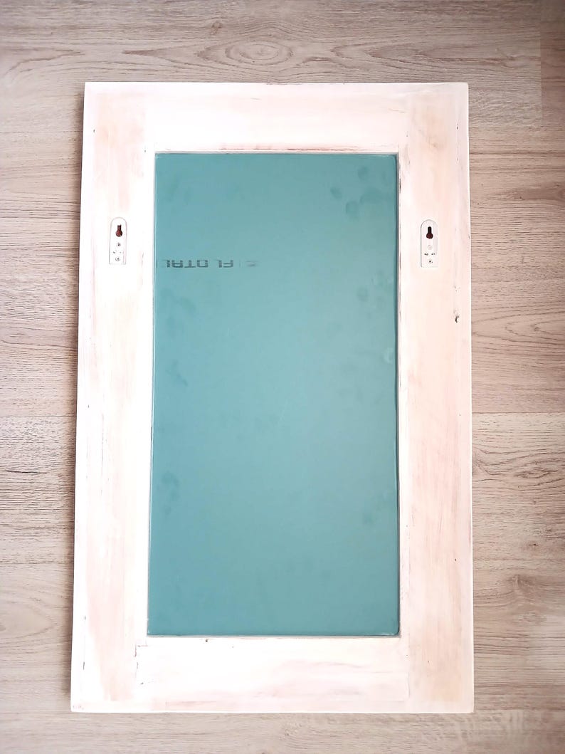 May include: White wooden frame with a light blue mirror inside. The frame has two small white hooks on the back. The mirror has the text "F1 0781" printed on it.