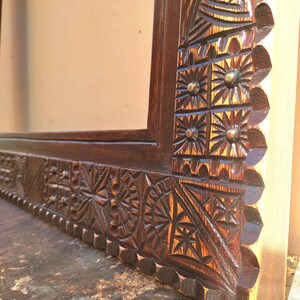 May include: Close-up of a dark brown wooden frame with intricate, hand-carved geometric and floral patterns. The frame's corner features detailed starburst and sun-like designs, with small, round metal accents. The wood grain is visible.