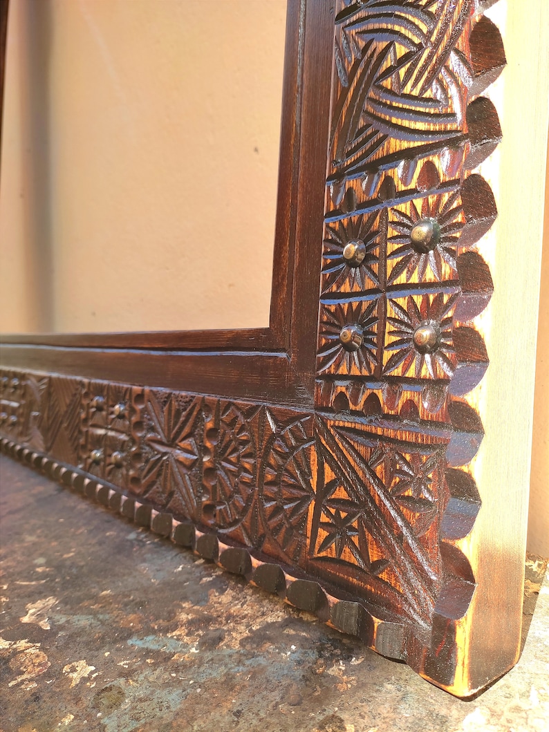May include: Close-up of a dark brown wooden frame with intricate, hand-carved geometric and floral patterns. The frame's corner features detailed starburst and sun-like designs, with small, round metal accents. The wood grain is visible.
