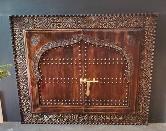 May include: A dark brown wooden door with intricate carvings and a brass latch. The door is framed by a decorative border with a repeating pattern.