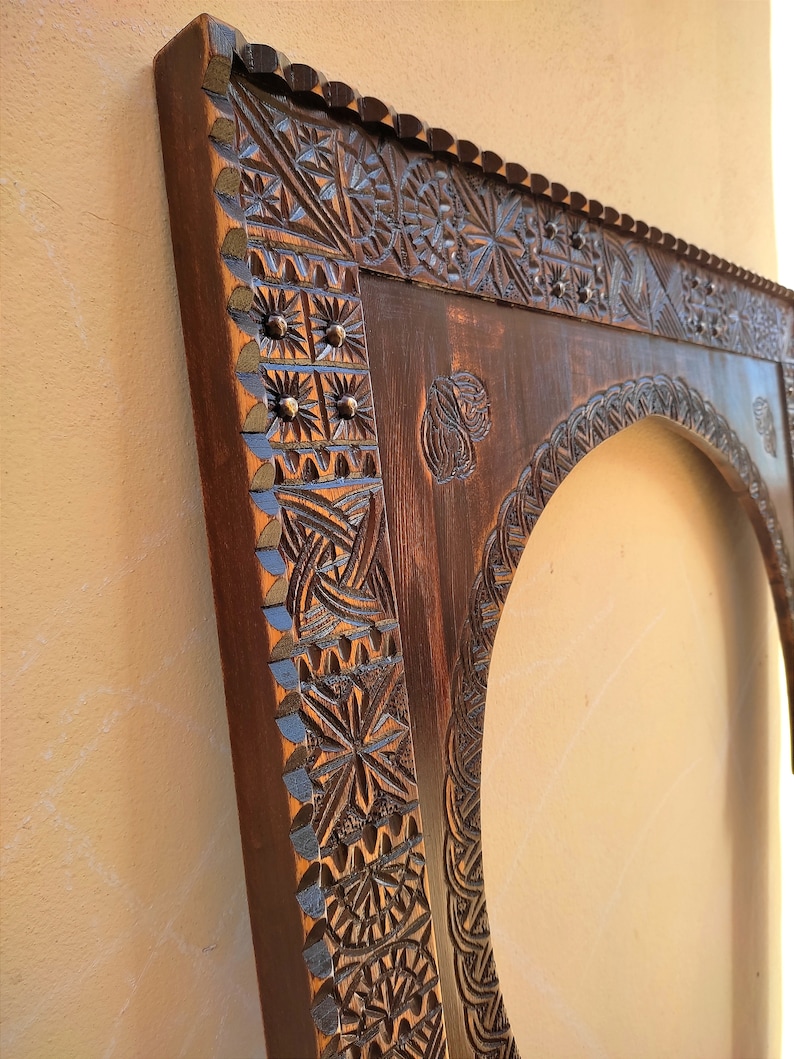 May include: A dark brown, intricately carved wooden frame with an arched opening. The frame features detailed geometric and floral patterns, with small metal studs. The wood has a rich, warm tone, and the carving is highlighted with a touch of blue paint.