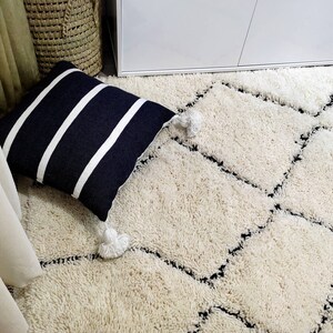 May include: A white shag rug with a black diamond pattern. The rug is in a room with a white cabinet and a black and white striped pillow.