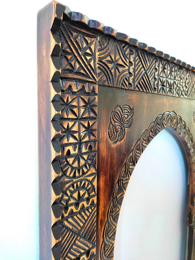 May include: A wooden frame with intricate carvings in a dark brown colour. The frame has an arched opening and is perfect for a mirror or artwork.