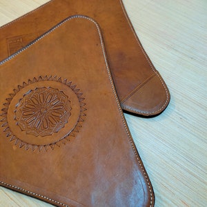 May include: Two brown leather triangular coasters with decorative stitching around the edges. One coaster features a circular design with intricate details.