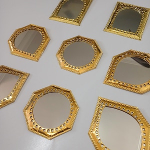 May include: An assortment of decorative gold-colored mirrors in various geometric shapes, including octagons, rectangles, and arched designs. Each mirror has a detailed, textured frame with a repeating pattern of small holes. The mirrors are arranged on a light gray surface.
