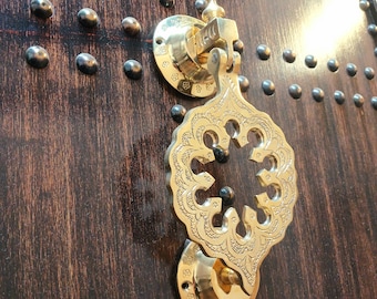 May include: Close-up of a decorative brass door knocker with an ornate, openwork design. The knocker is mounted on a dark brown wooden door, accented with numerous small, dark metal studs.