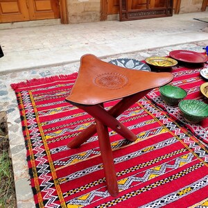 May include: A brown leather Moroccan pouf with intricate embossed designs sits on a red and yellow patterned rug. The pouf has a triangular shape and is supported by three wooden legs.