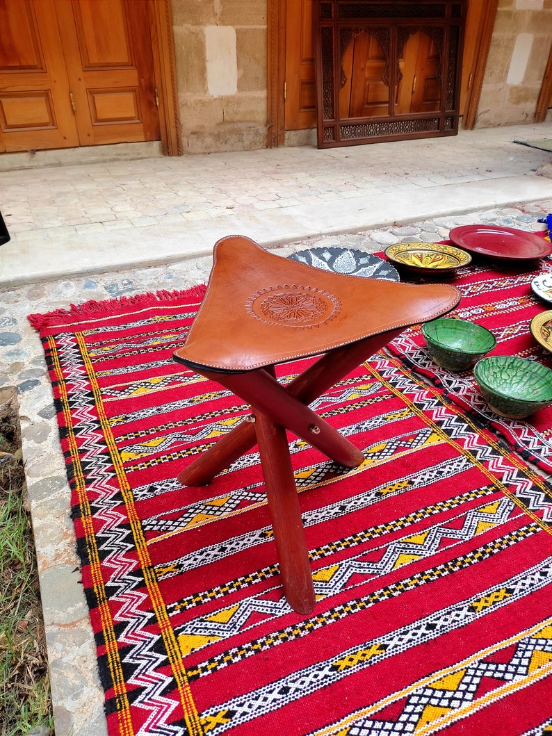 May include: A brown leather Moroccan pouf with intricate embossed designs sits on a red and yellow patterned rug. The pouf has a triangular shape and is supported by three wooden legs.