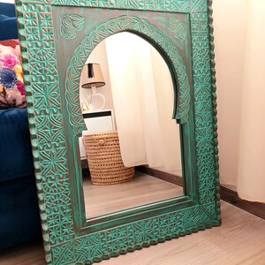 May include: A teal green wooden mirror with an arched top and intricate carved details. The mirror is framed with a decorative border and has a distressed finish.