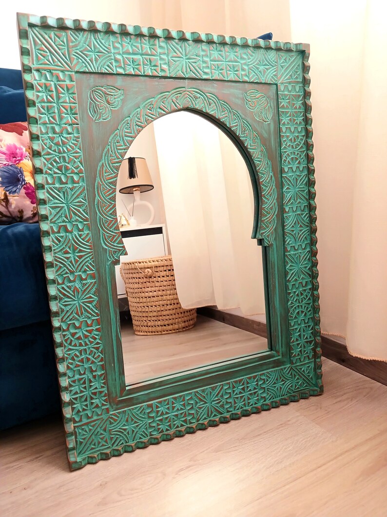 May include: A teal green wooden mirror with an arched top and intricate carved details. The mirror is framed with a decorative border and has a distressed finish.