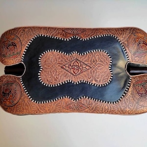 May include: A brown leather ottoman with intricate carvings and a dark blue center panel. The ottoman has a unique shape with two rounded ends.